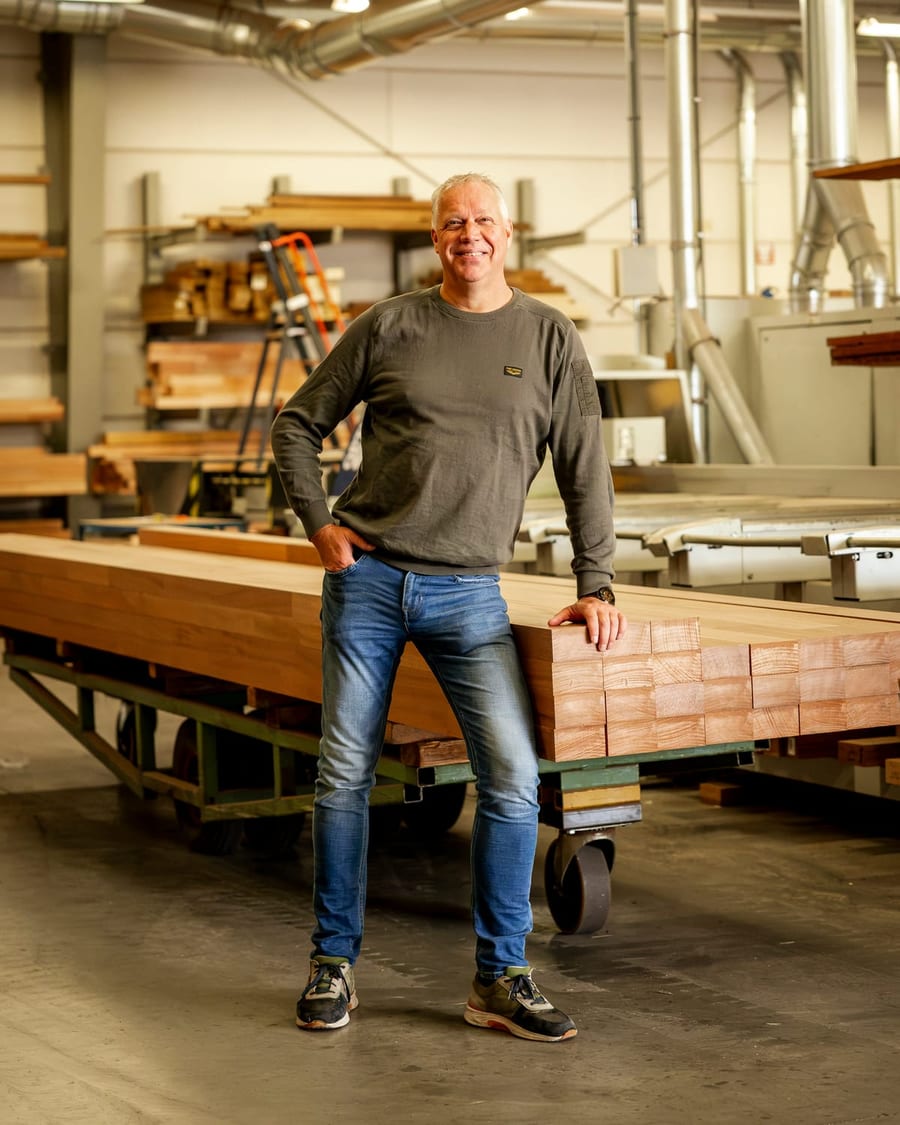 Jan Vermeer staat lachend in de fabriek, met een hand op een stapel geschaafde balken