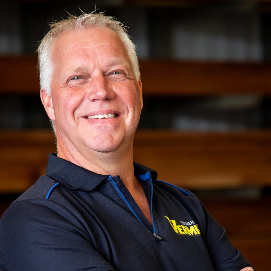Jan Vermeer, lachend met de armen over elkaar voor de houtopslag